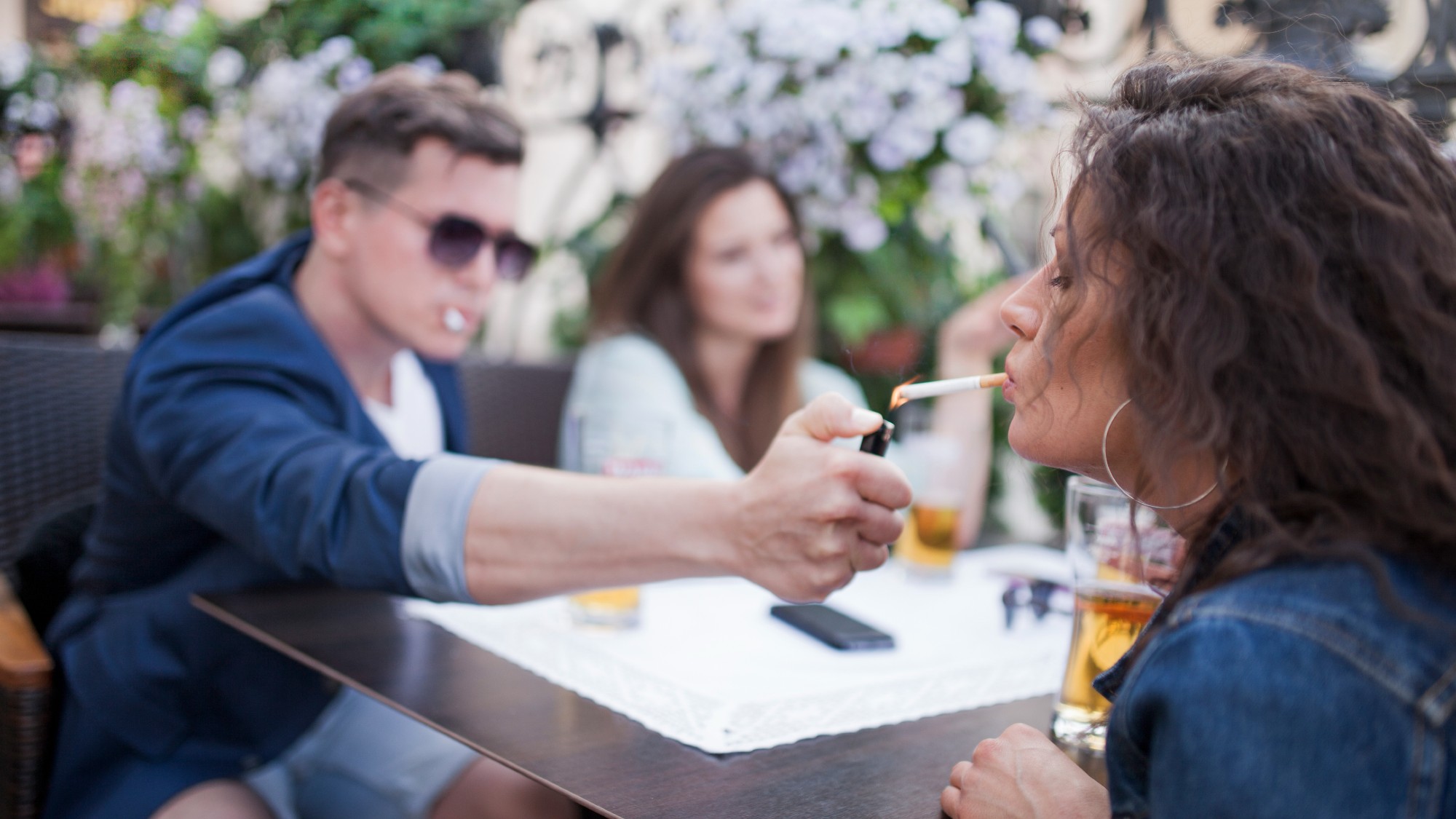 Group of young people drinking beer in a beer garden, one man leaning across the table to light a female companion&#039;s cigarette.