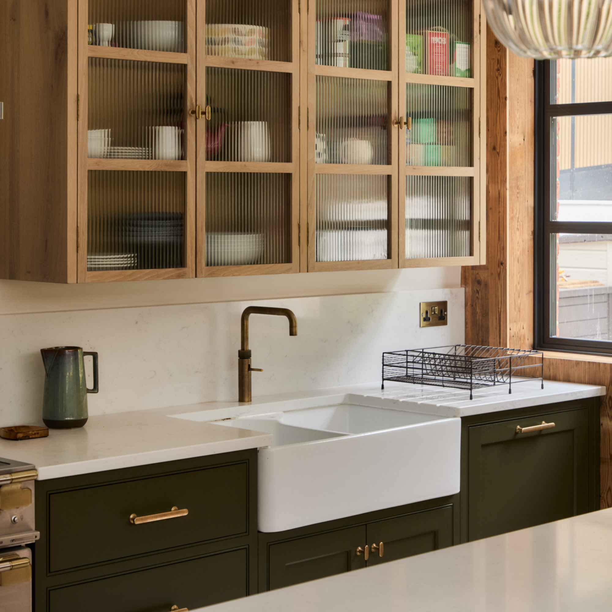 kitchen with dark green painted units, white work tops and sink, natural wood wall unit with fluted glass doors