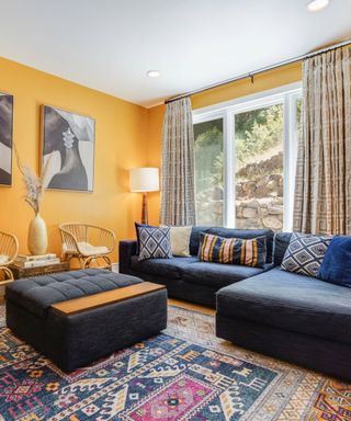 A yellow living room with curtains, a window, a blue couch and ottoman, and a colorful rug