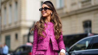 Woman in a matching pink tweed suit and black glasses, looking a way from the camera with wavy hair over her shoulder