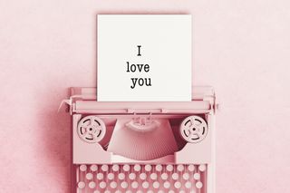 A pink typewriter displaying a piece of paper with the words 'i love you' written, on a light pink background.