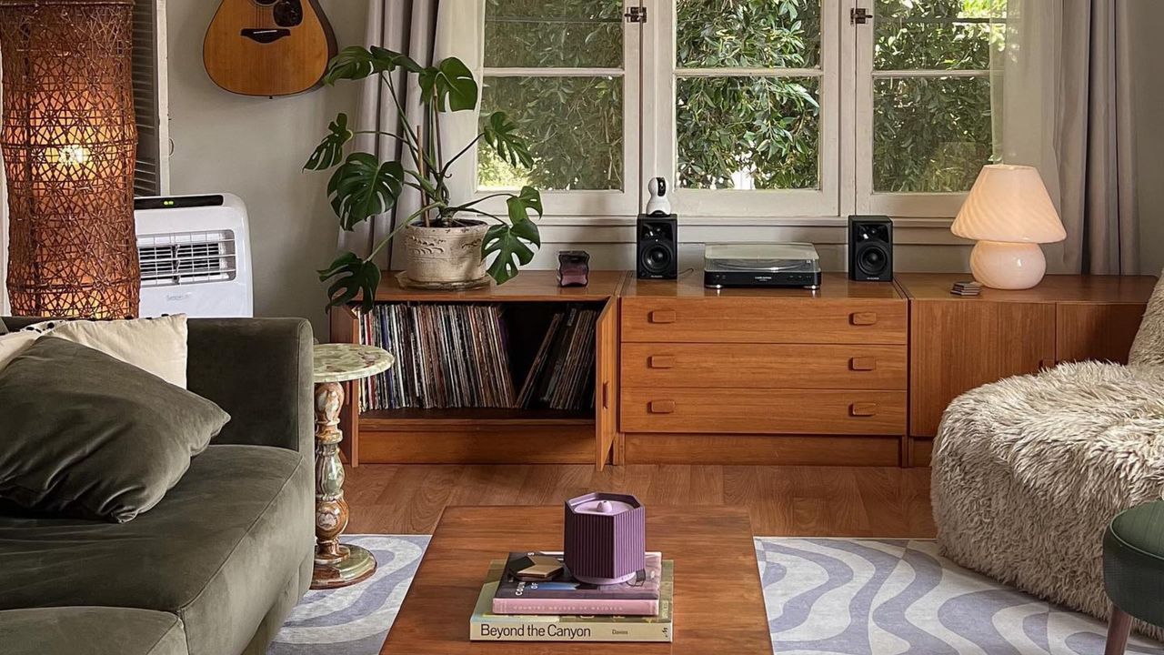 An image of a tidy small living room with hardwood floors, an olive-green velvet couch, a fluffy cream armchair, a wooden coffee table, and a purple and white wavy rug with natural accents and lighting