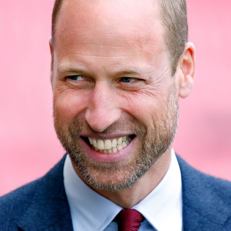 Prince William laughs, and has a rugged beard, while wearing a blue suit with a white shirt and red tie 