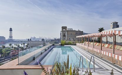 View of the roof top pool and row of red and white lounge beds at Soho House. There is also a view of nearby buildings and trees under a blue sky