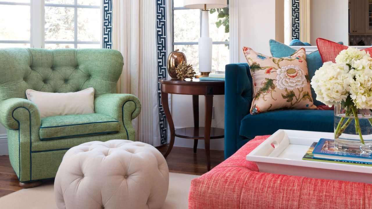 A living room with a pistachio green chair, a white tufted storage ottoman, a navy blue couch and coral coffee table, and two large windows behind them