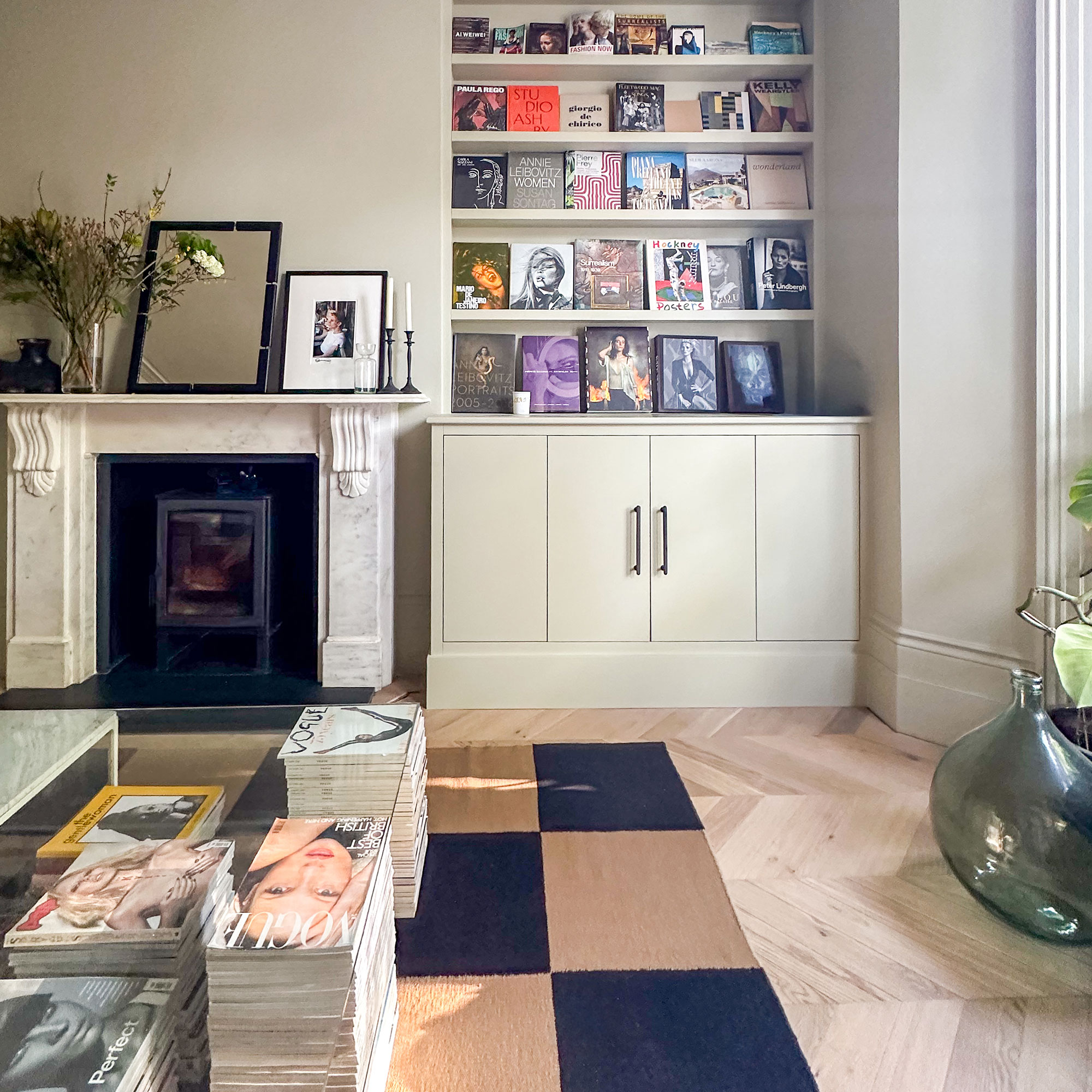 Living room with chequered rug, glass table propped up with magazines, pale green built-in storage holding display of coffee table books