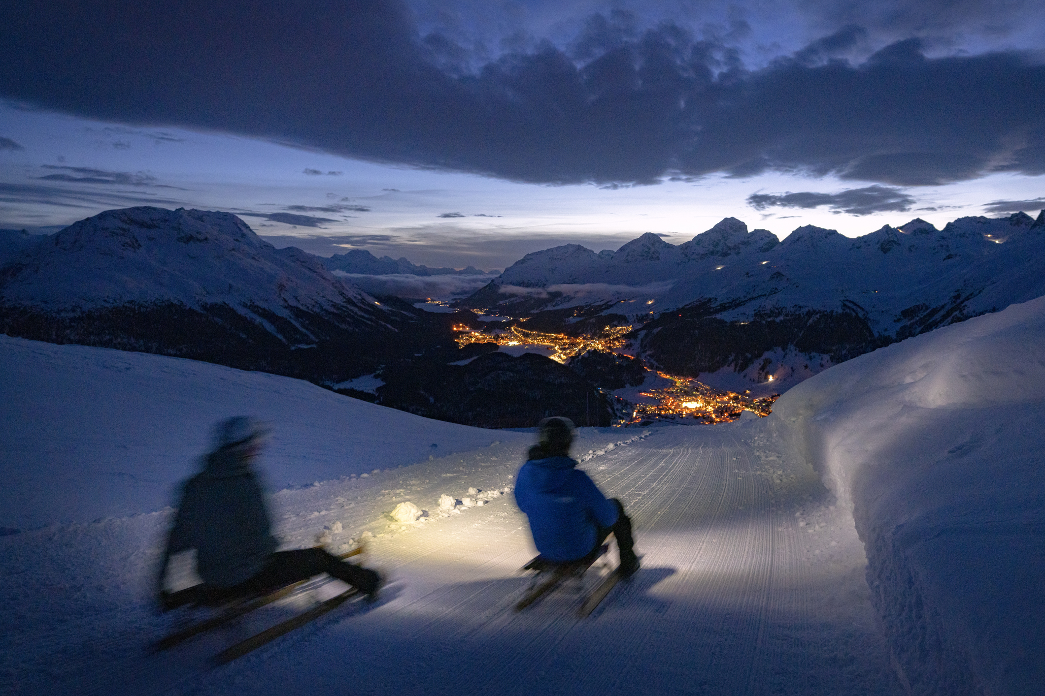 Tourists enjoying sledding on snowy mountains at moonlight, Muottas Muragl, St. Moritz, Graubunden canton, Engadine, Switzerland.