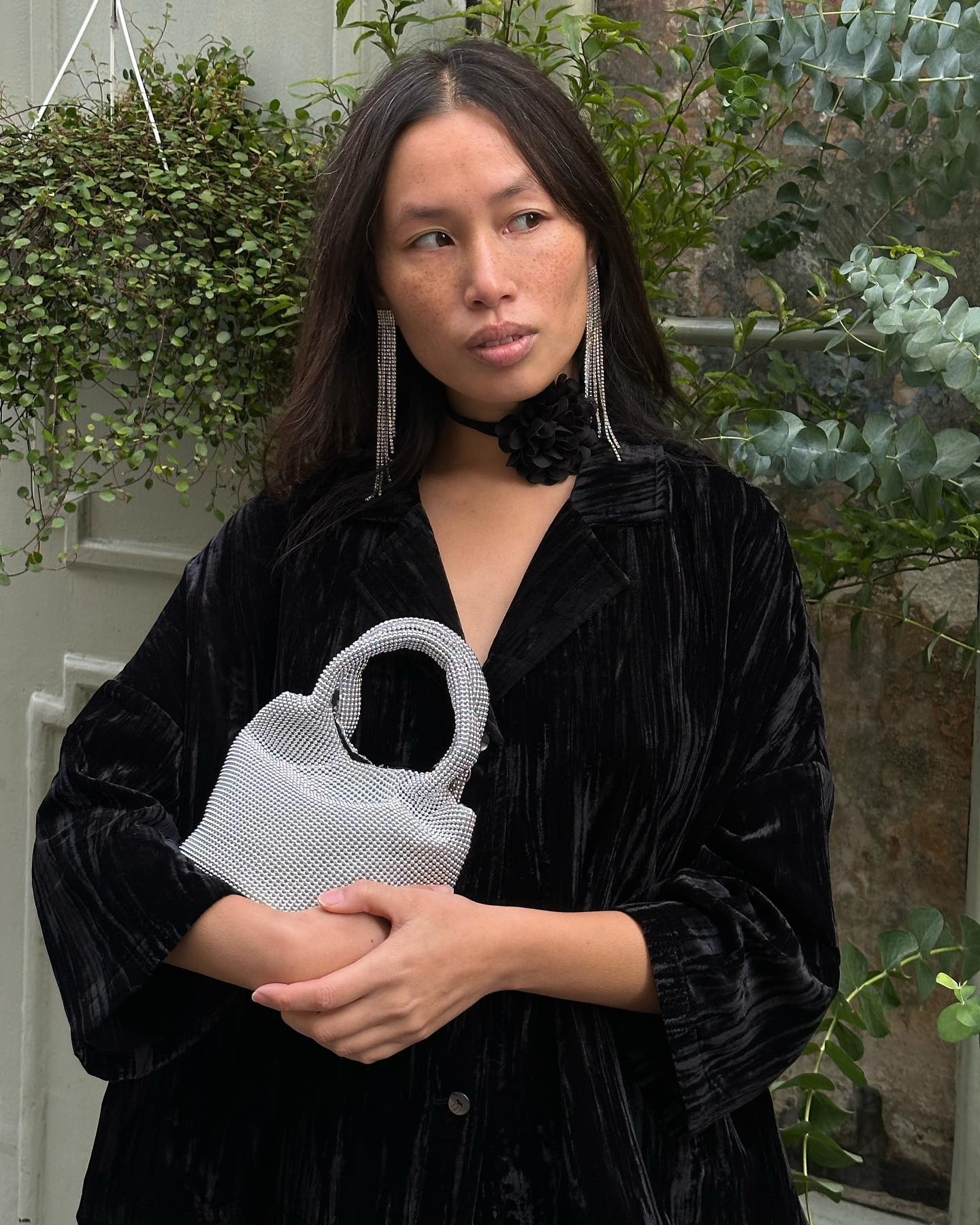 Paris based style influencer Anne-Victoire Lefevre wearing long rhinestone earrings, a black flower choker, a black velvet jacket, and a Parfois silver beaded mini bag
