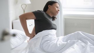 A woman in a brown t-shirt massages her painful lower back because she's sleeping on a memory foam mattress that's too soft for her and she should switch to a hybrid bed