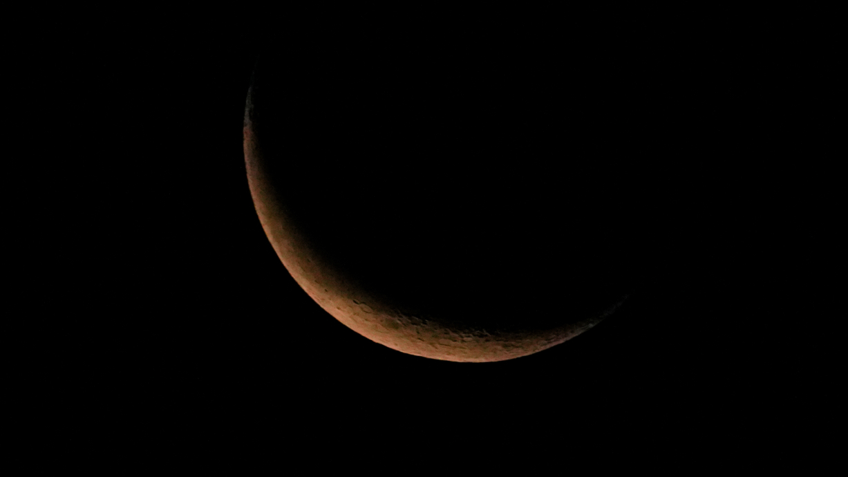 Photo of an orange, thin, waning crescent moon against a black sky.