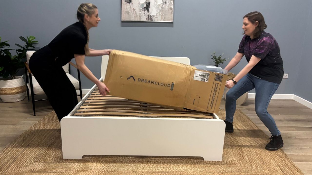 Two mattress testes unbox the DreamCloud Luxe Memory Foam mattress, taking it out of its box, cutting it out of its plastic wrapping and placing it on a white bed frame
