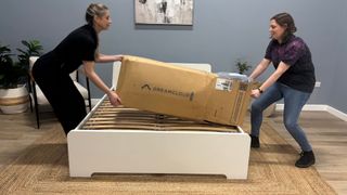 Two mattress testes unbox the DreamCloud Luxe Memory Foam mattress, taking it out of its box, cutting it out of its plastic wrapping and placing it on a white bed frame