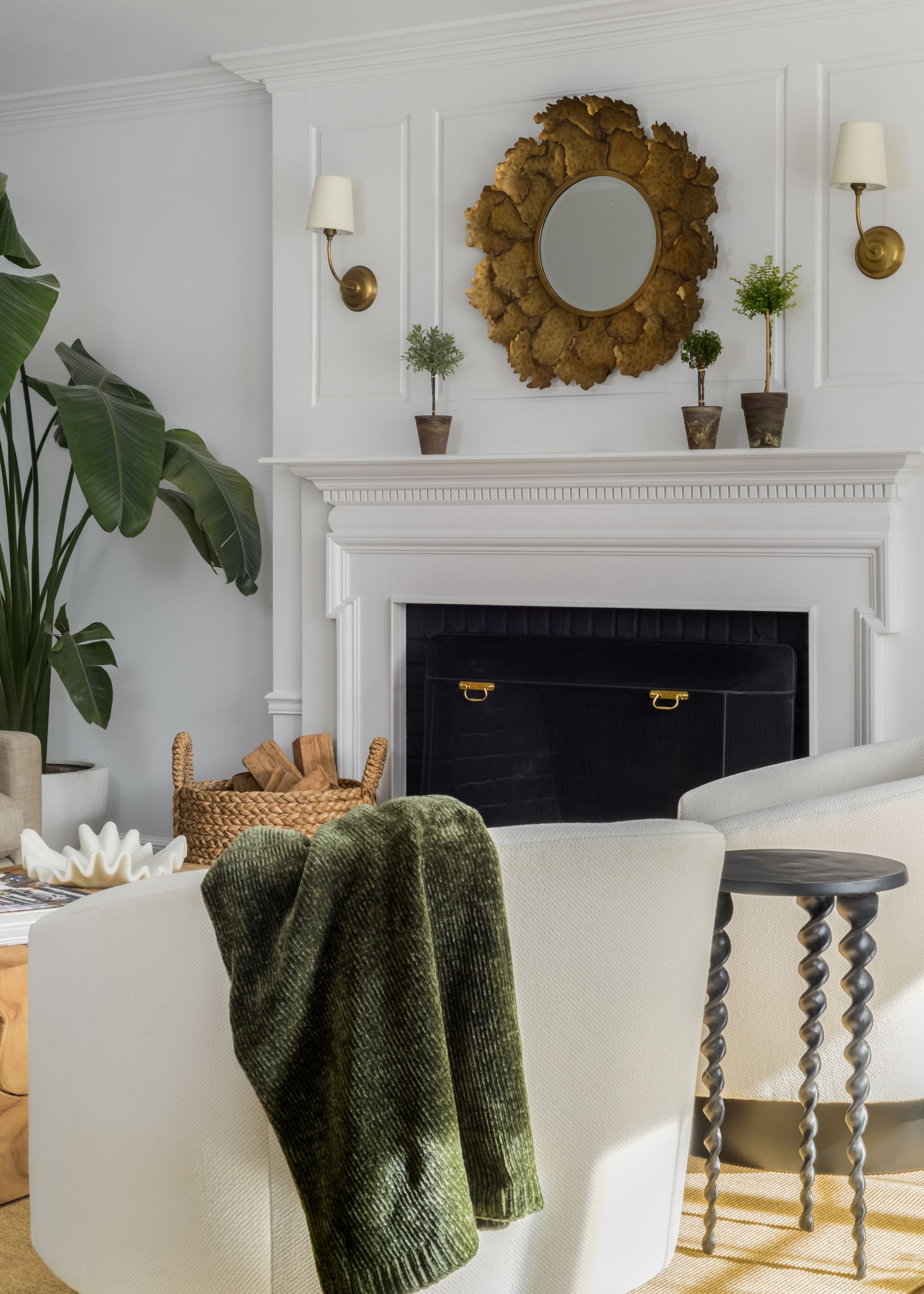 A living room with white walls and a white fireplace, wall lights above the fireplace, and a gold circular mirror.