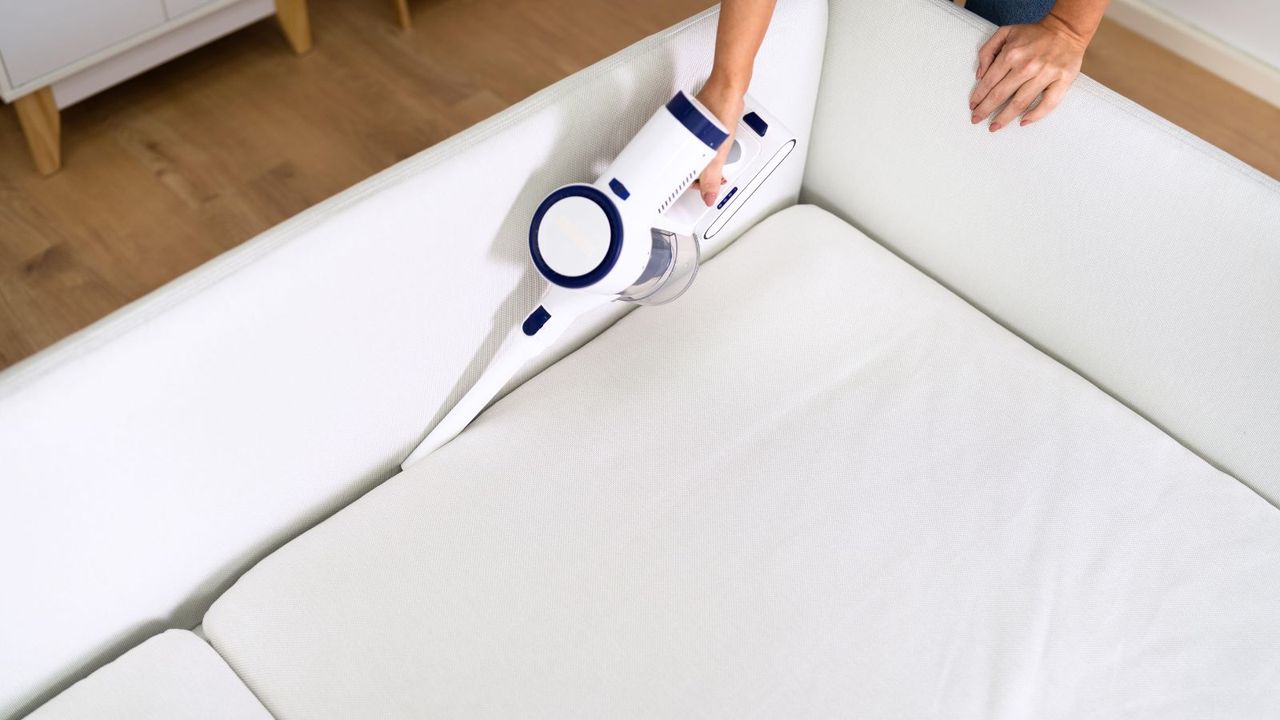 Someone using a hand held vacuum to clean the crevice of a white couch