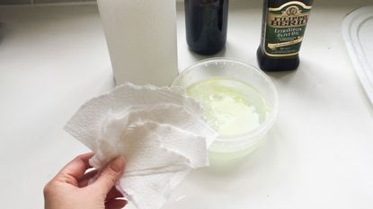 a hand holding pieces of torn up kitchen cloth, with a kitchen roll, bowl full of water, and olive oil too – all the essential for the tiktok cleaning hack for oily plastic food containers
