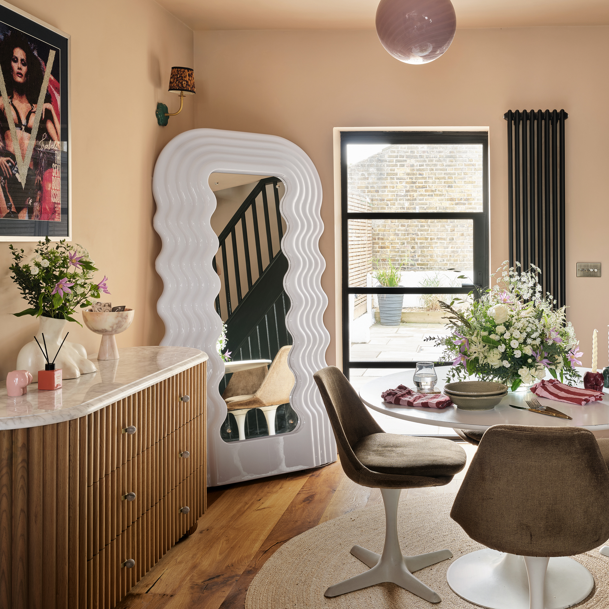 an open plan dining room with a Tulip table and matching upholstered chairs, a chest of drawers with a wood fluted front and marble top and a large UltraFragola designer mirror in the corner beside a patio door