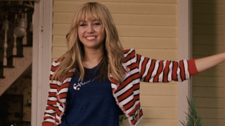 Miley Cyrus smiling in front of a house as Hannah Montana in Hannah Montana: The Movie