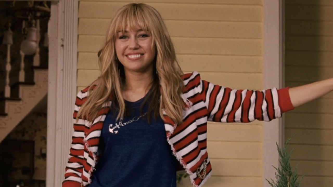 Miley Cyrus smiling in front of a house as Hannah Montana in Hannah Montana: The Movie