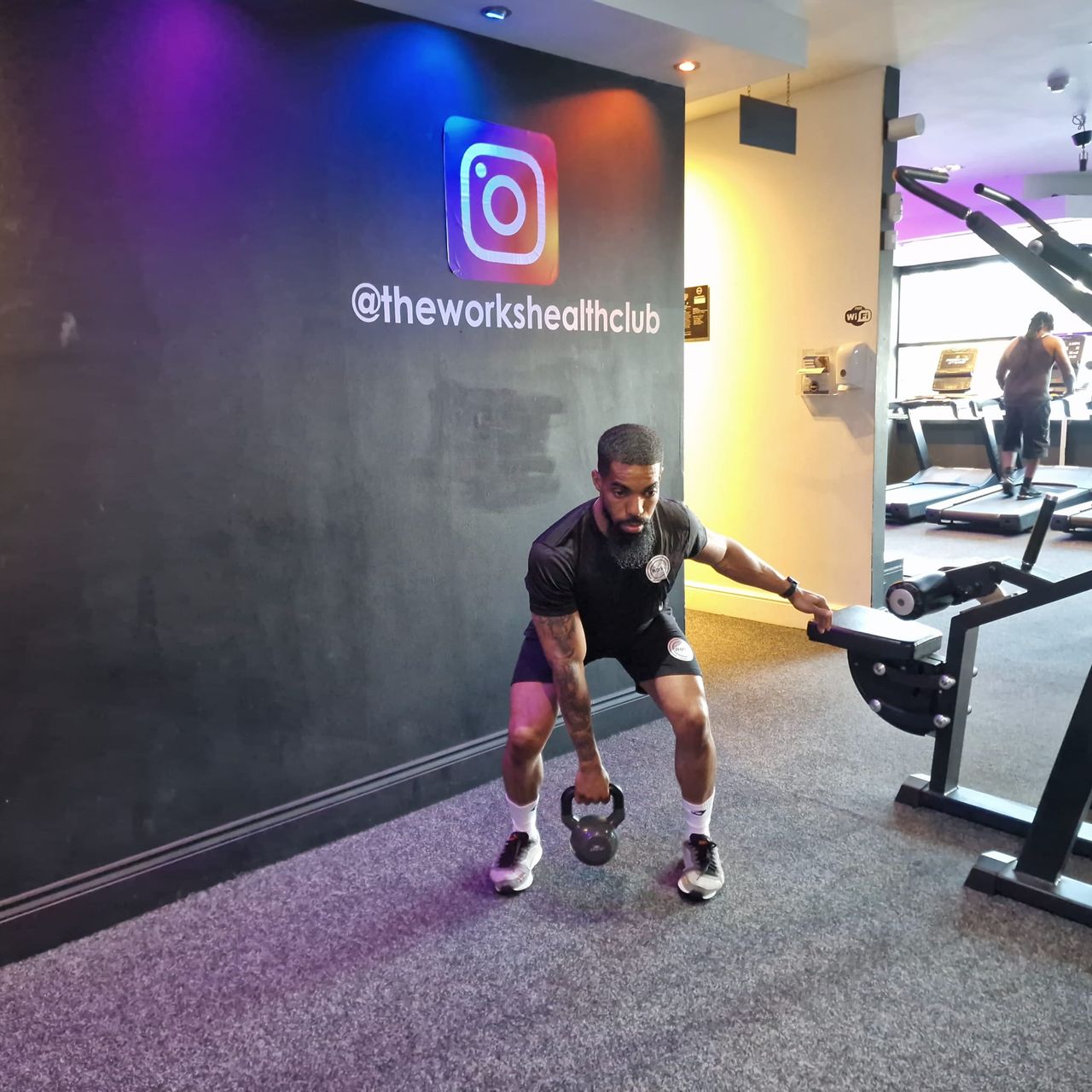 Renel Scarlett performs a kettlebell snatch in The Works Health Club gym. He is squatting down, bending at both his knees and hips, with one arm held behind him and the other extended downwards to grasp a kettlebell. Behind him we see a person on a treadmill.