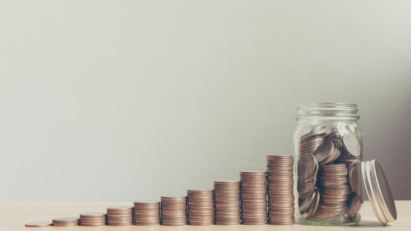 Coins are stacked in piles that get higher alongside a jar of coins