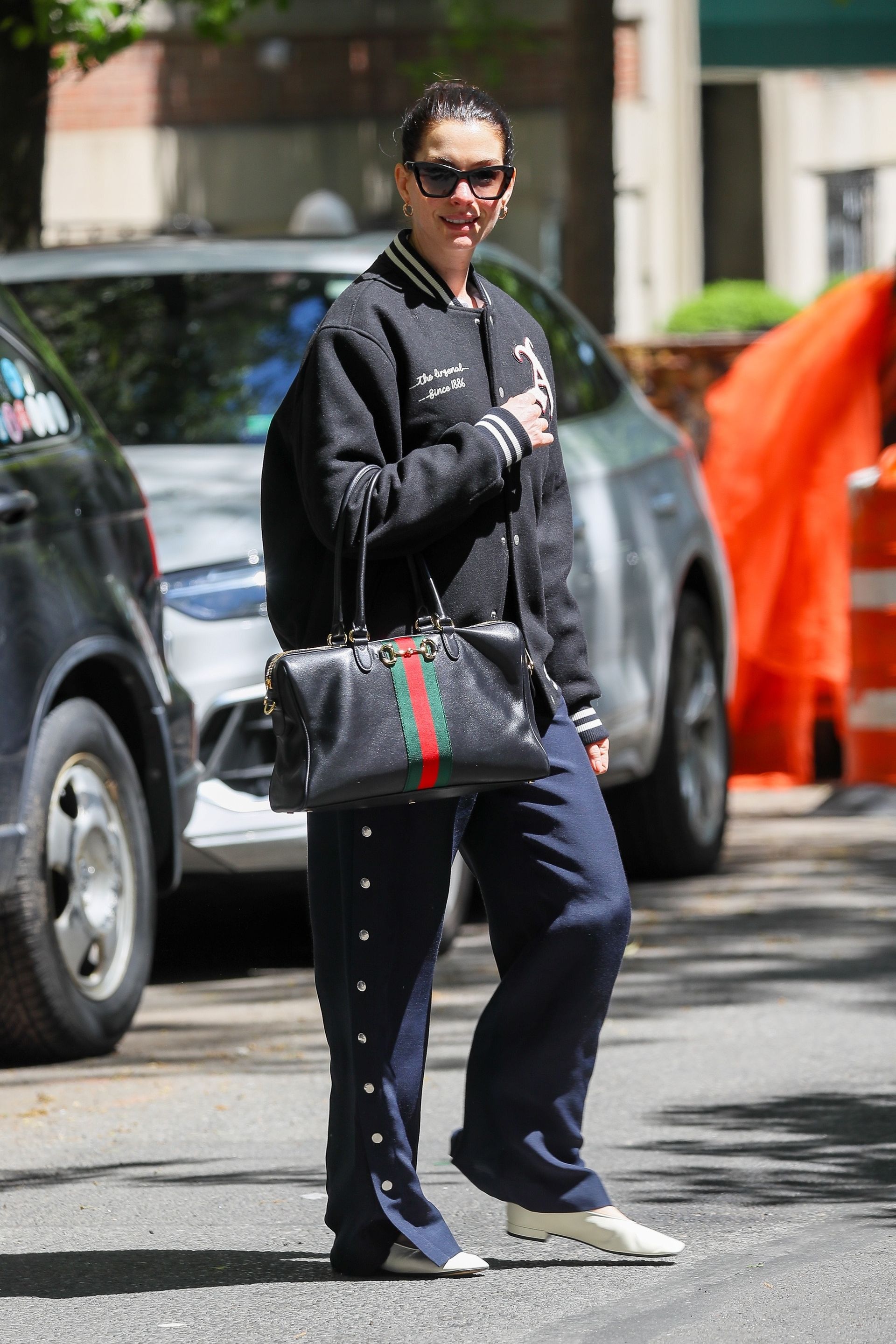 Anne hathaway wears a bomber jacket, navy trousers, and white loafers with gucci borsetto bag.