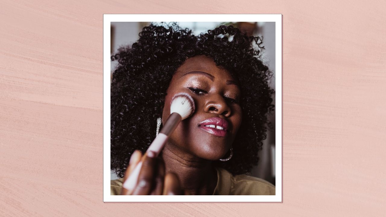 A woman with curl hair is pictured applying blush to the top of her cheeks with a fluffy brush/ in a peach-pink template