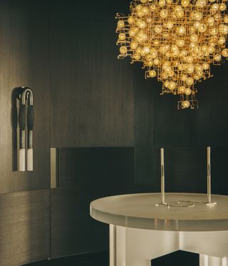 A round glass island with a multi-bulb chandelier hanging above. Colour theme is predominantly dark grey and brown.