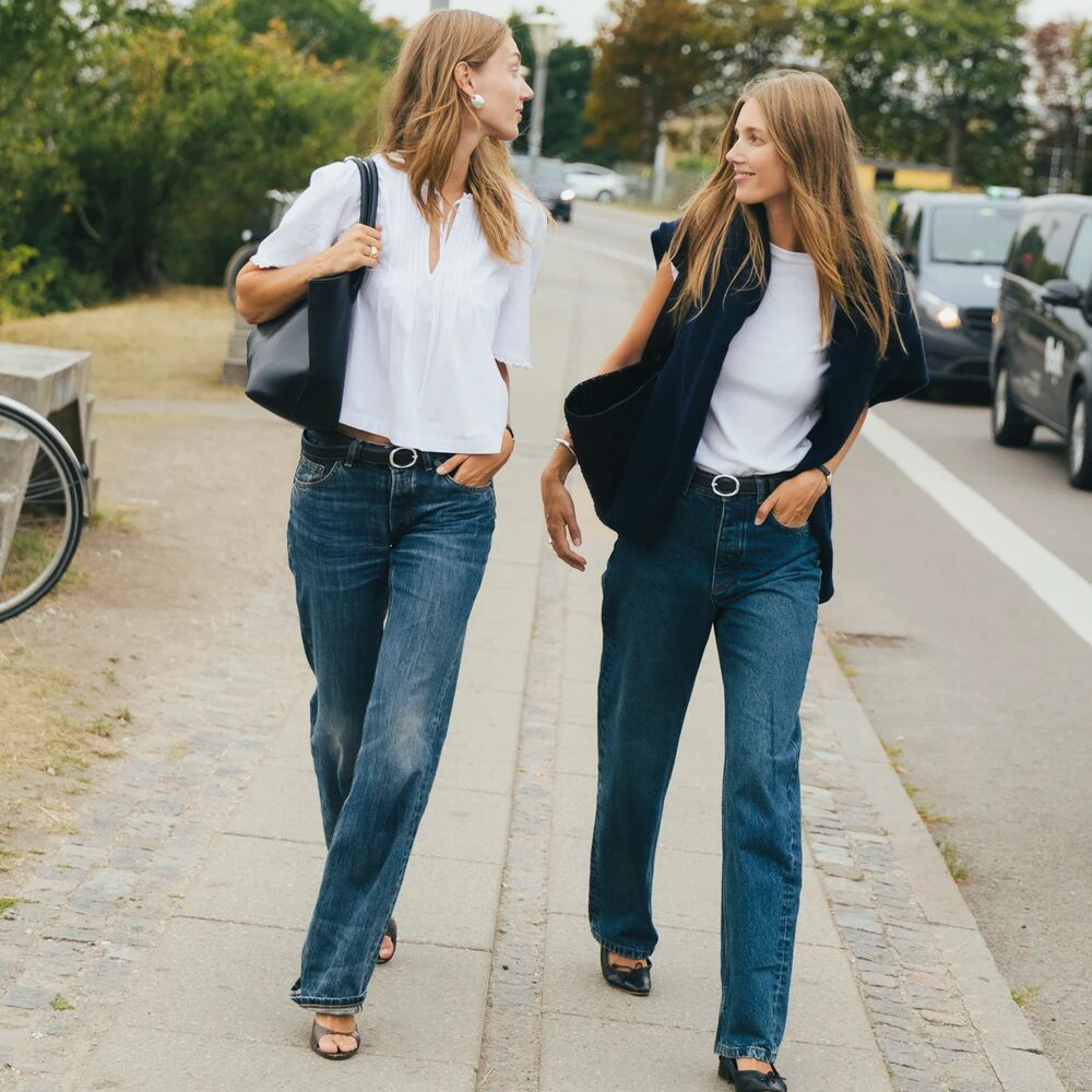 Two women are walking and talking in Copenhagen while posing. One wears a white t-shirt, flats, and jeans. Another wears white button-down, jean, and heels.