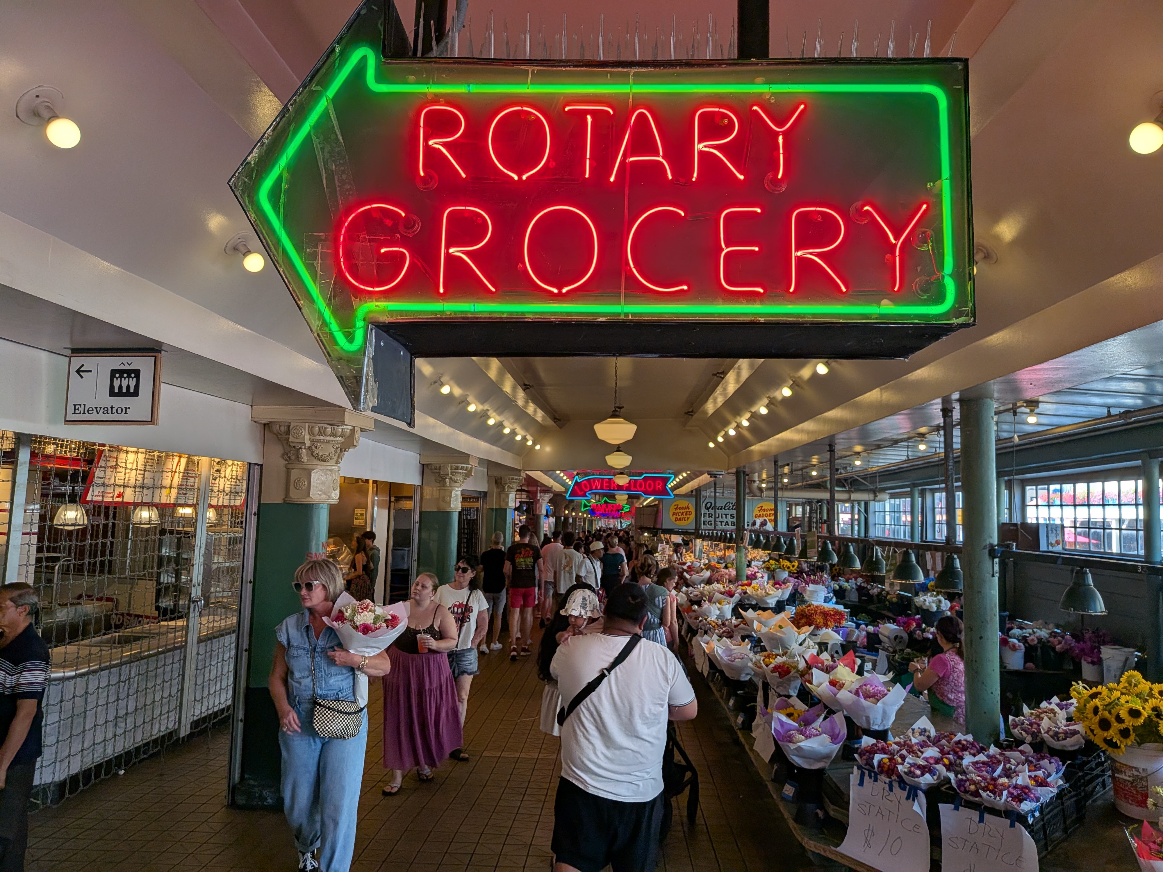 A neon Rotary grocery sign above a indoor walkway with flowers on one side and people on another