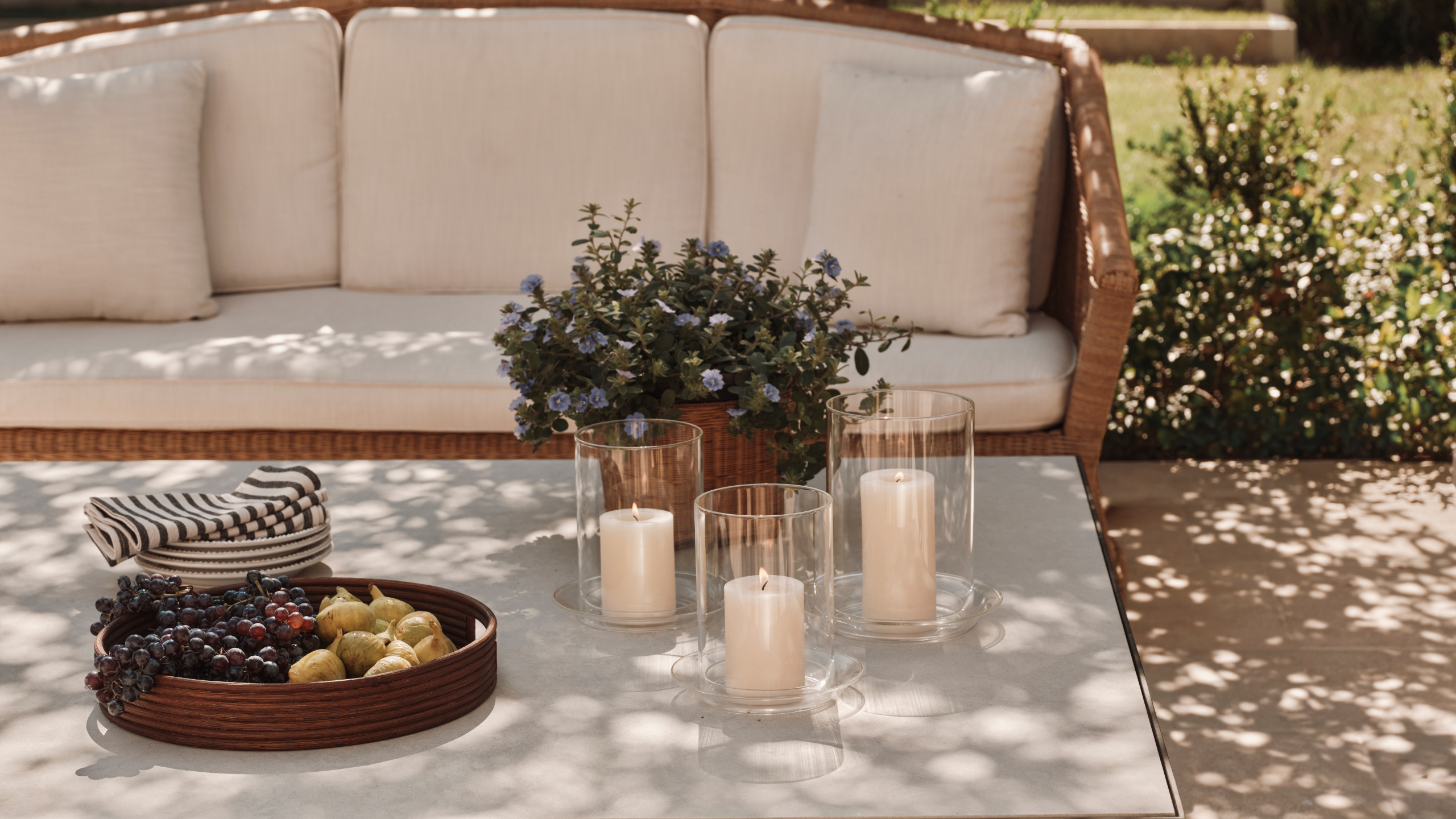 Sunny California outdoor living room featuring a wicker outdoor sofa and stone coffee table topped with a rattan circular tray, candle hurricanes, and a basket of wildflowers.