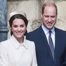 Princess Kate wears a white coat and matching hat while her husband wears a navy suit