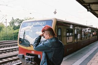 A passenger with red mo hawk and blue denim jacket, standing on the platform of a train station , with a train passing by