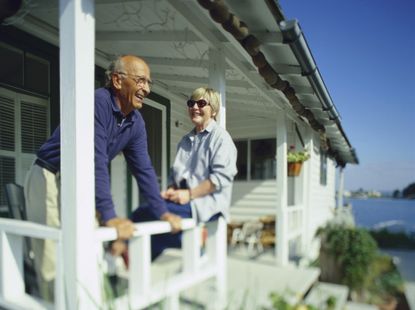 An older couple smiles as they relax on the porch of a beach house. The mood is casual and it is a sunny day.