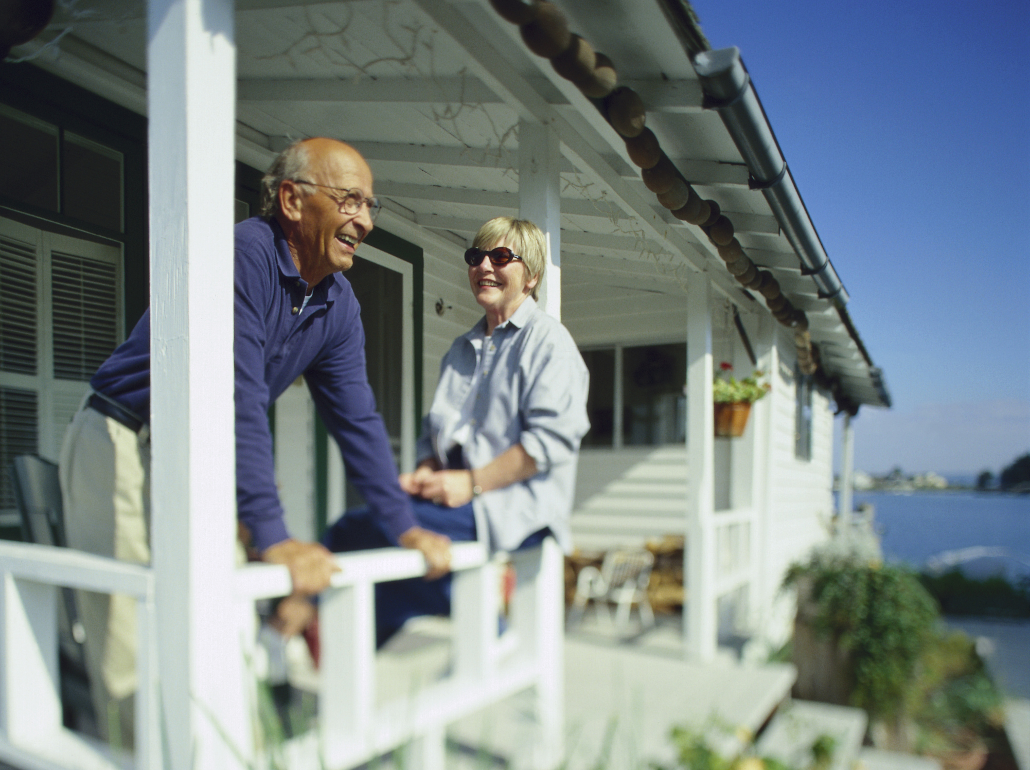 An older couple smiles as they relax on the porch of a beach house. The mood is casual and it is a sunny day.