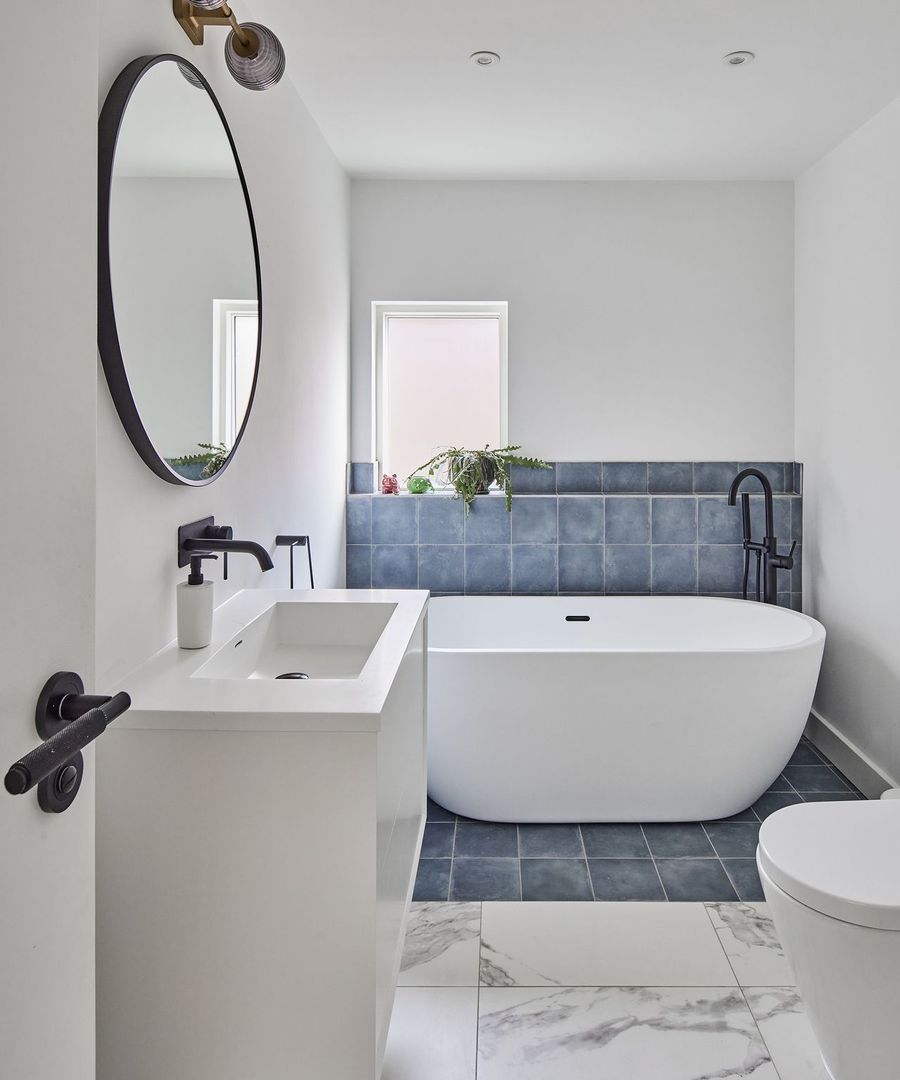 A bathroom with freestanding bath standing on blue tiles. The remaining floor tiles are a large marble effect. There is a rectangular sink with black taps and a circular mirror