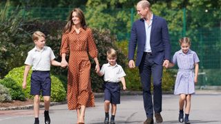 Princess Kate and Prince William holding hands with Prince George, Prince Louis and Princess Charlotte, all wearing navy school uniforms, walking into school with trees behind them