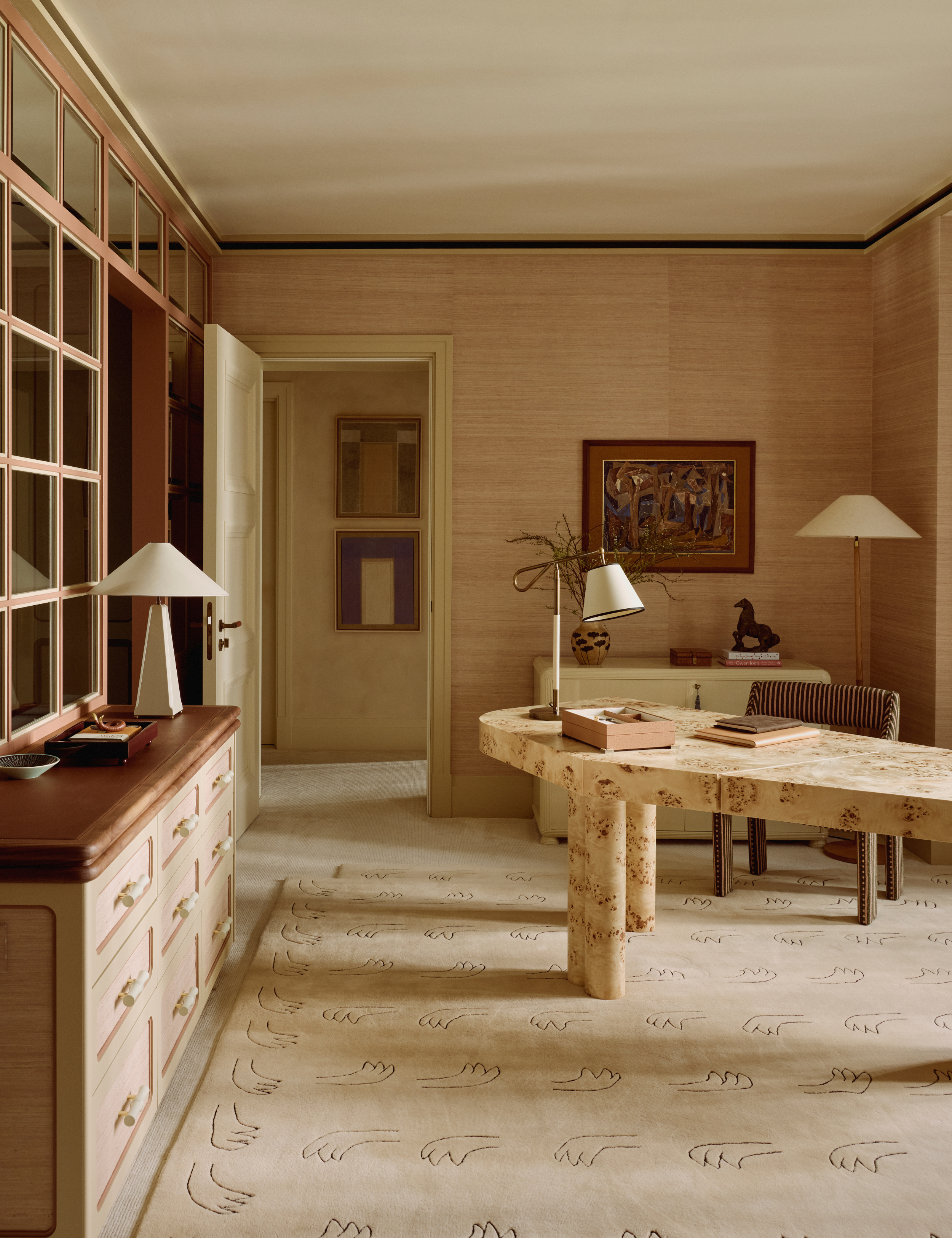 Neutral office space featuring an asymmetrical burl wood desk, linen-shaded table lamps, wall art, textured wallpaper, a figural motif beige rug, and a striped office chair.