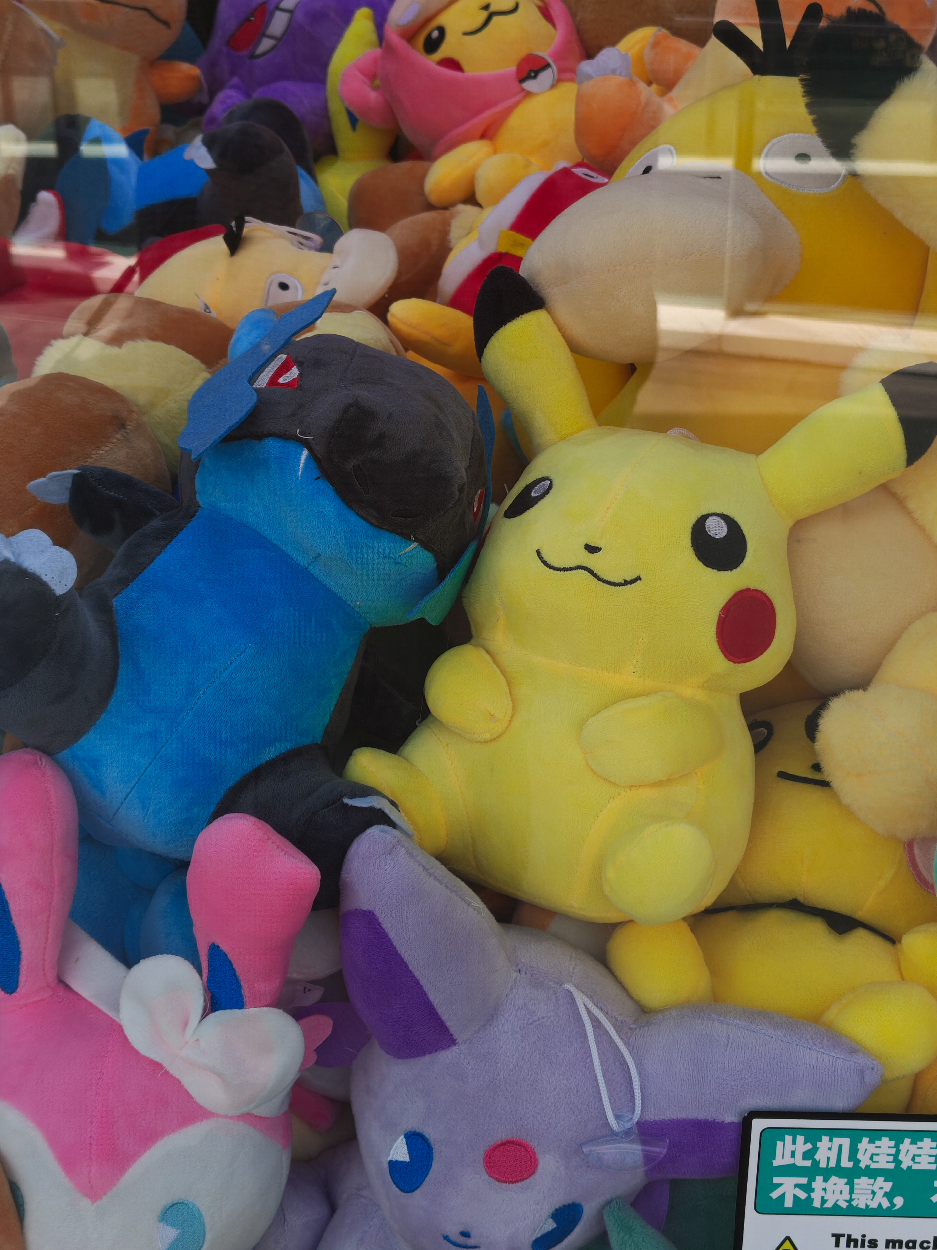 A close-up shot of a variety of plush toys inside a claw machine. A bright yellow Pikachu plush is the central focus, surrounded by a blue and black Pokemon-style plush, a purple Eevee-like plush, and several others. The glass of the machine shows slight reflections of the surrounding environment.