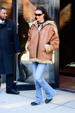 A photo of Bella Hadid wearing a brown leather shearling-lined jacket, light wash flare jeans, and a black graphic tee topped with a gold coin charm necklace in New York City.