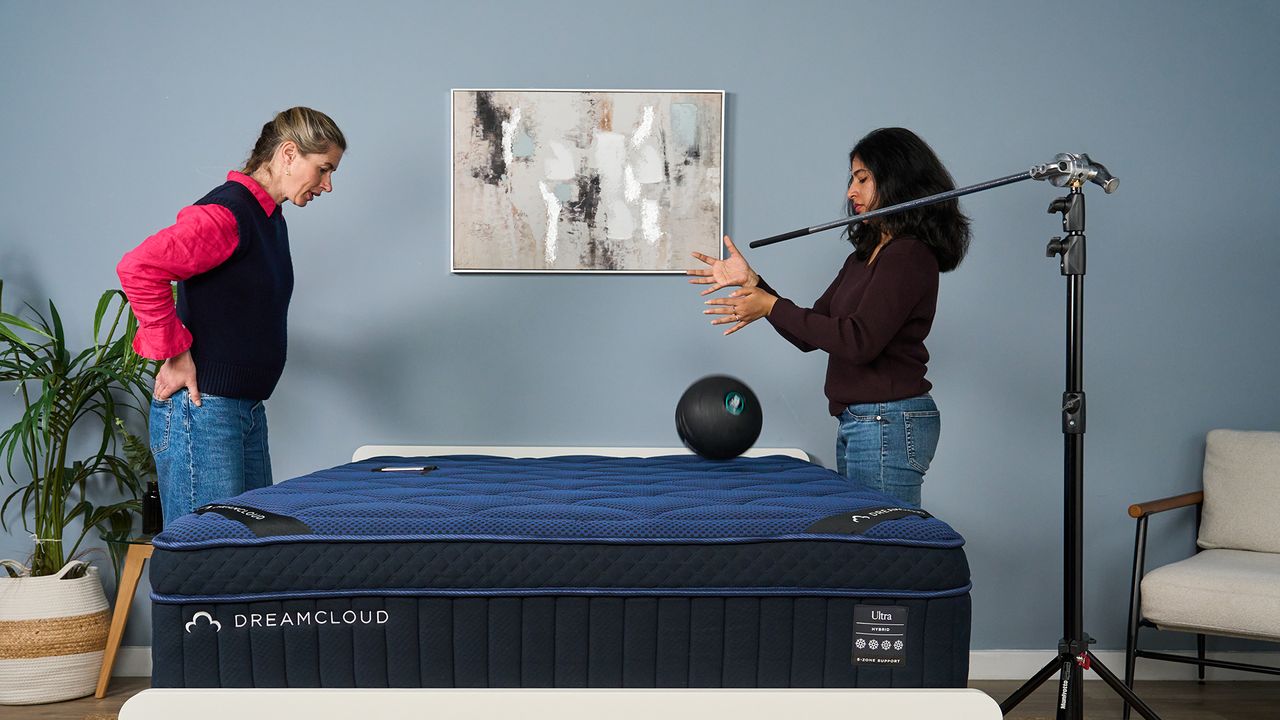 Two mattress testers stand above a blue mattress, with one dropping a 5kg slam ball onto its surface while another records its reading on her phone. This helps measure motion isolation.