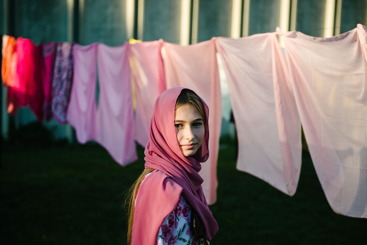 A girl in a pink headscarf stands in front of a line of hanging pink and patterned fabrics, with a softly lit background