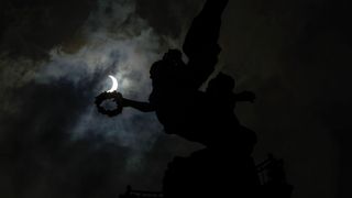 A partial eclipse peers through cloudy skies over an angel statue