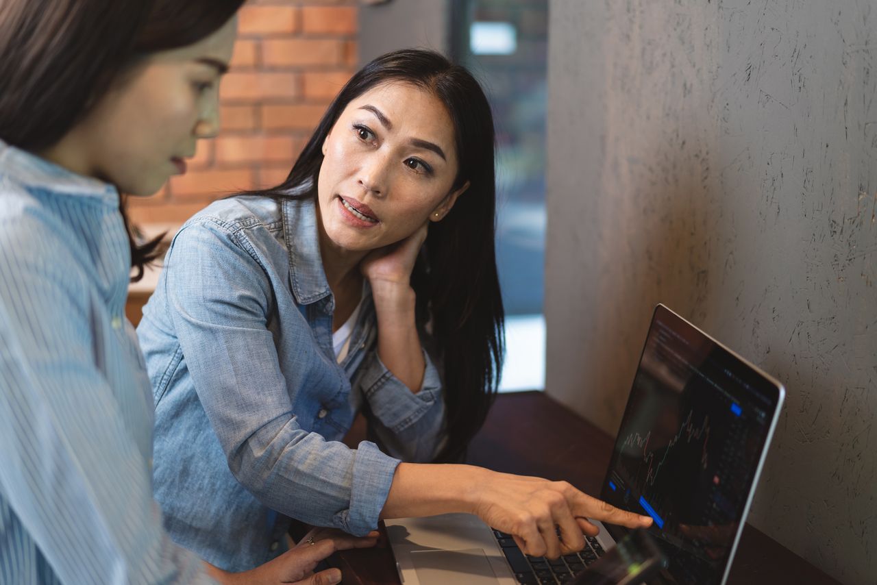 One woman teaching another how to buy stocks by looking at a stock chart on a laptop