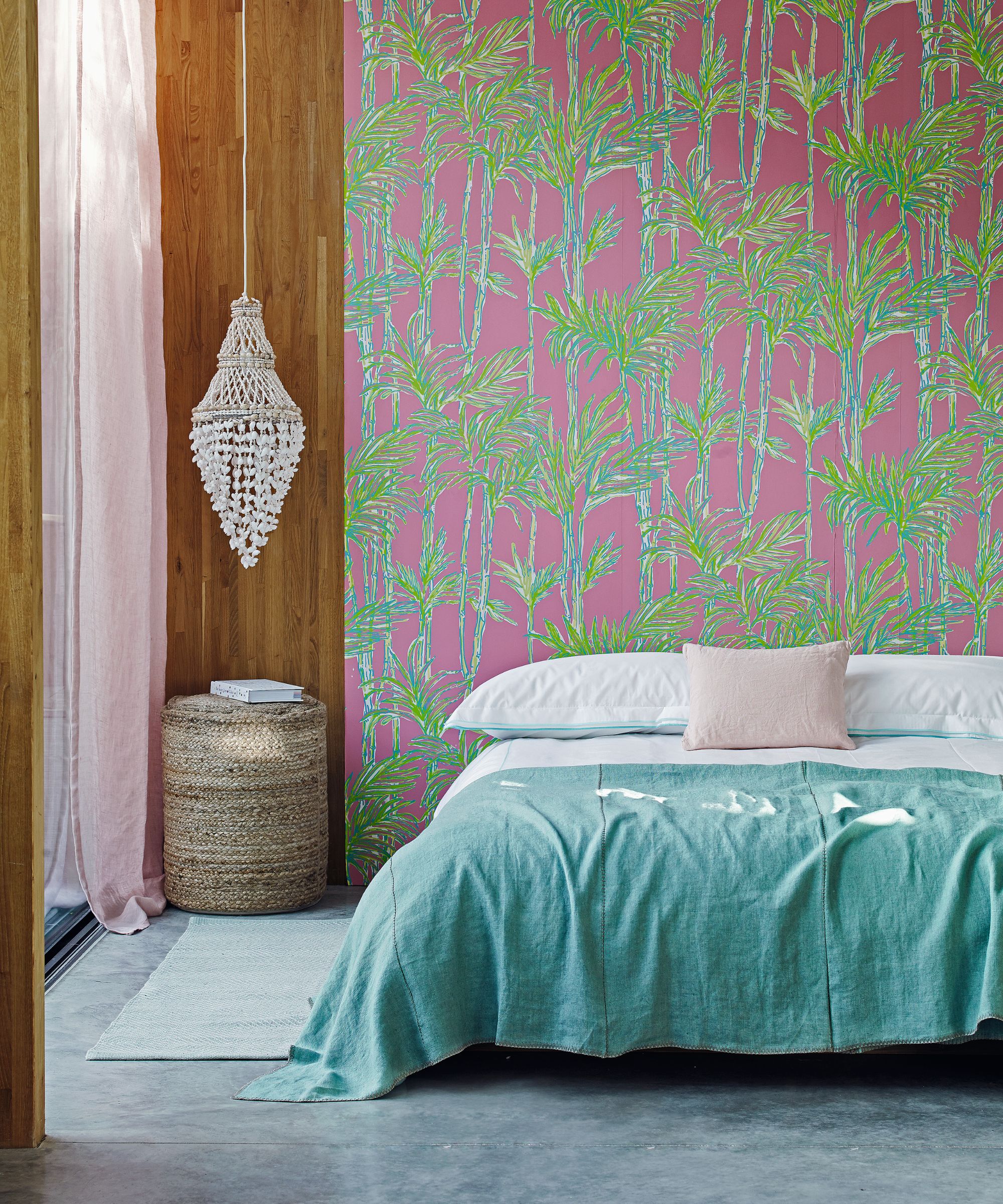 A colorful bedroom with a pink and green patterned statement wall, wood panelling, a macrame hanging, rattan stool, and a low bed with blue bedding.