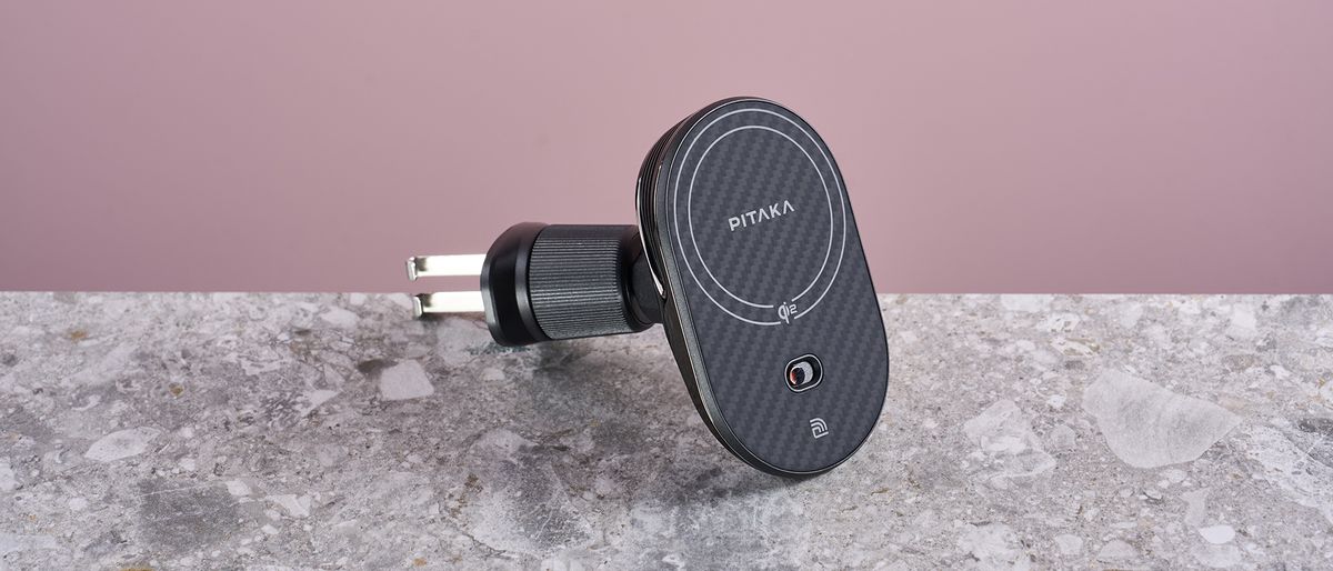 The Pitaka MagEZ Car Mount Pro 2 Qi2 sits on a stone-effect surface with a pink background. The view is of the front profile at a slight angle, revealing the collar fasteners and the metal attachment hooks.