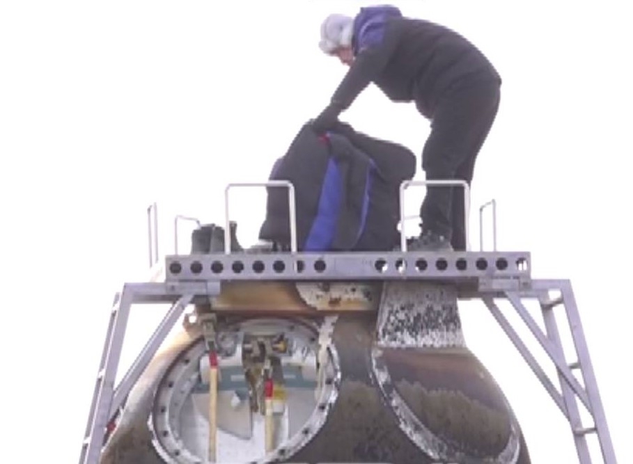 A person wearing all black stands on top of a metal space capsule and bends down to look into its open top hatch