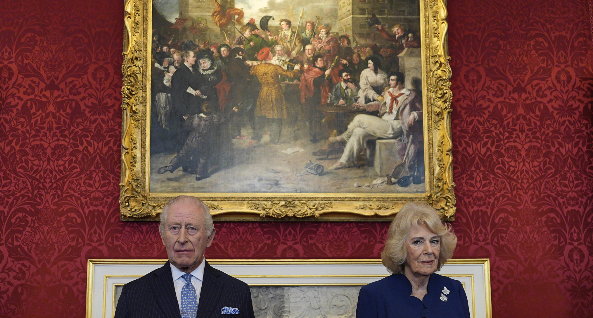 King Charles and Queen Camilla standing in front of a painting on a red wall with serious expressions