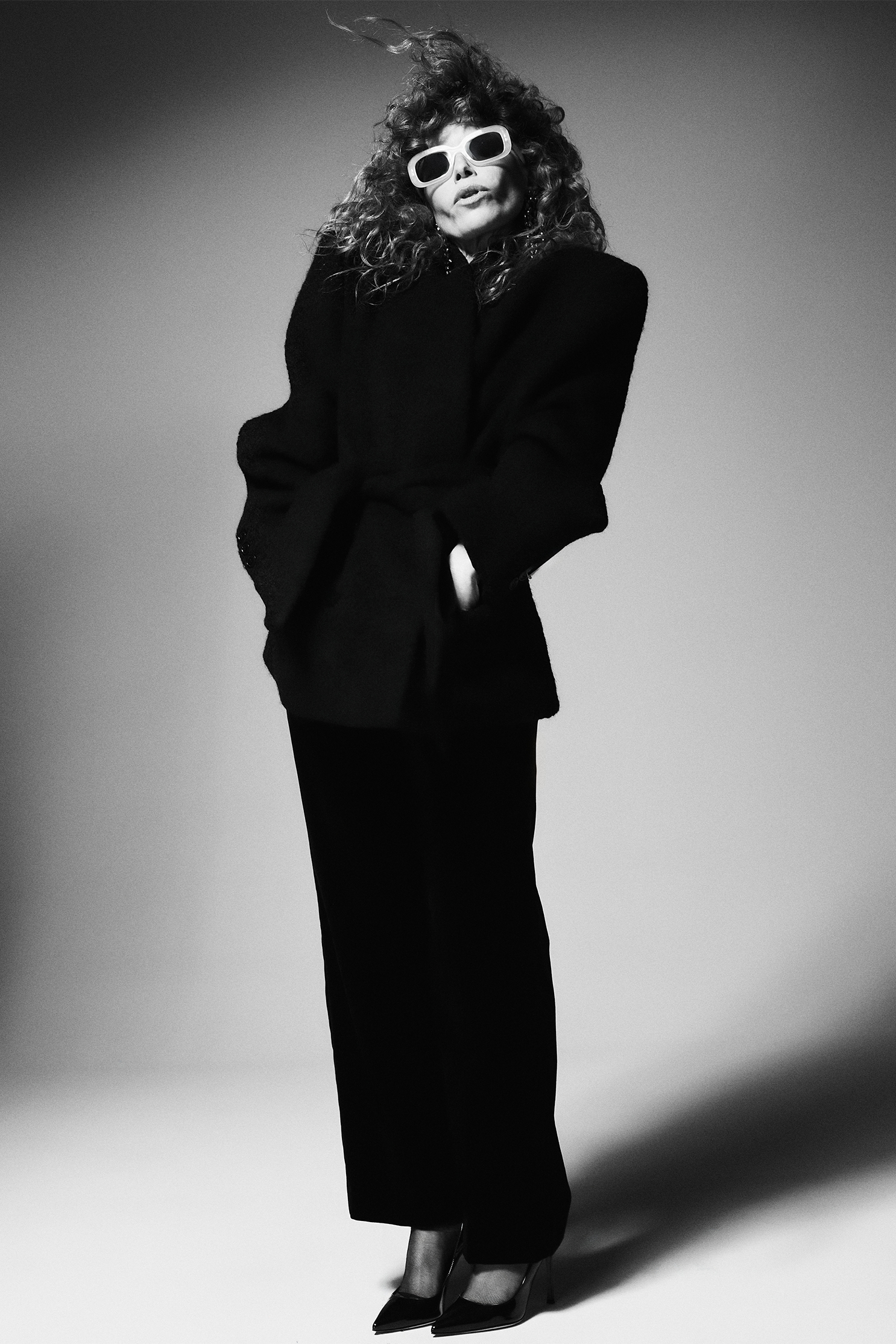 Natasha Lyonne photographed by Andrew Arthur for Who What Wear. She is wearing white-frame sunglasses and a black suit with her hands in her pockets.