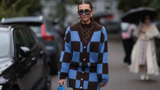 Lina Uhrig seen wearing Corlin Eyewear black sunglasses with blue transparent lenses, gold earrings, Gestuz brown / black triangle print pattern blouse, Stine Goya brown / blue checked knit wool cardigan, matching Stine Goya rown / blue checked knit wool short skirt, Jacquemus bright blue leather Bambino bag, Cartier Love gold ring, outside Stine Goya, during the Copenhagen Fashion Week Spring/Summer 2024 on August 09, 2023 in Copenhagen, Denmark.