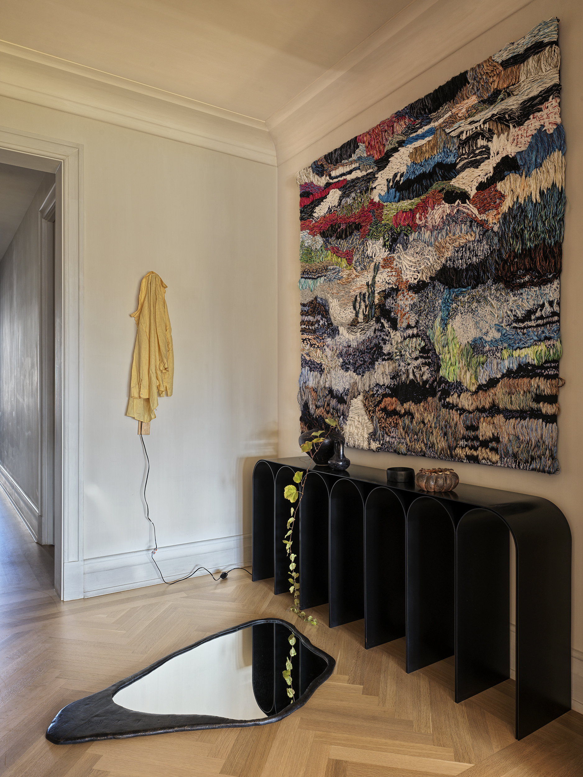 A bold tapestry hangs above a black modern console in an entryway with parquet wood flooring with a black framed abstract mirror lying on the floor and a yellow jacket hanging on the white wall